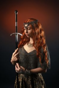 A woman with red hair wearing a chain-mail dress holding a sword up in her hands